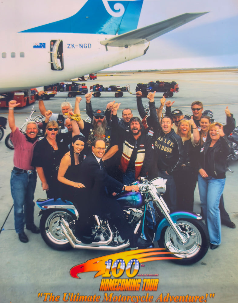 Melbourne Airport, 2003. A promotional photograph celebrating the Homecoming Tour’s partnership with Air New Zealand. Chris Beattie stands centre, Doug from Air New Zealand sits on a Harley Davidson, with "Mistress Alicia" riding pillion. A small group of tour members joined the tarmac photo shoot beside one of Air New Zealand’s aircraft.